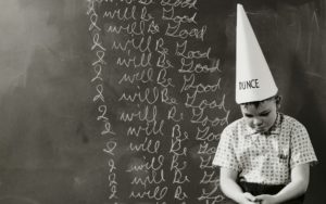 child sitting in front of chalkboard with dunce hat